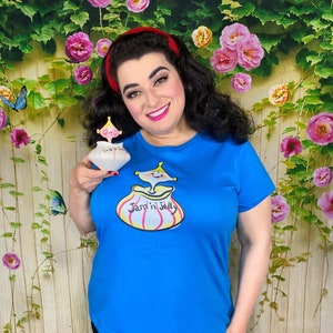 Puede incluir: Una mujer lleva una camiseta azul con una ilustración de dibujos animados de un tarro de mermelada con el texto "Jam'n Jelly". Está sosteniendo un pequeño tarro de cerámica con un diseño similar y el texto "Jam'n Jelly".