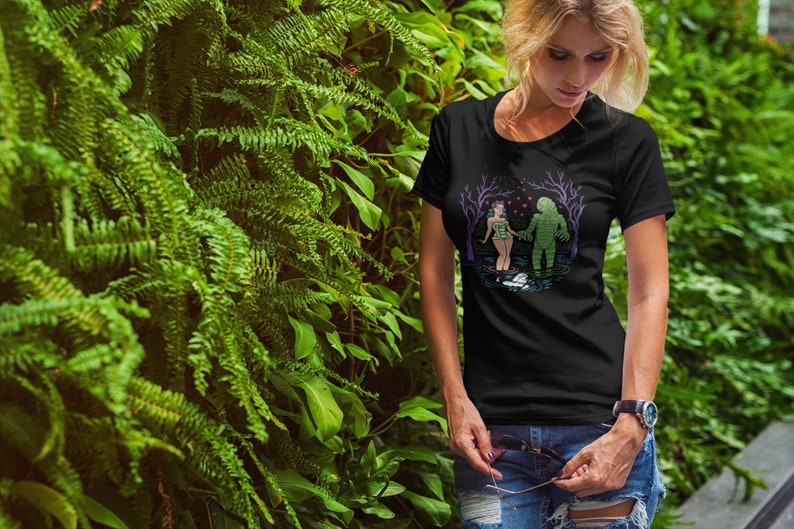 May include: Black t-shirt featuring a graphic of a swamp monster and a woman holding hands in a swamp. The design includes trees, hearts, and a small boat. The shirt is worn by a person in front of a green leafy background.