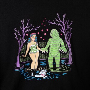 May include: Black graphic t-shirt featuring a cartoon illustration of a woman in a striped swimsuit holding hands with a green monster in a swamp. Pink hearts float above them, with a small dog swimming in the water. Purple trees frame the scene.