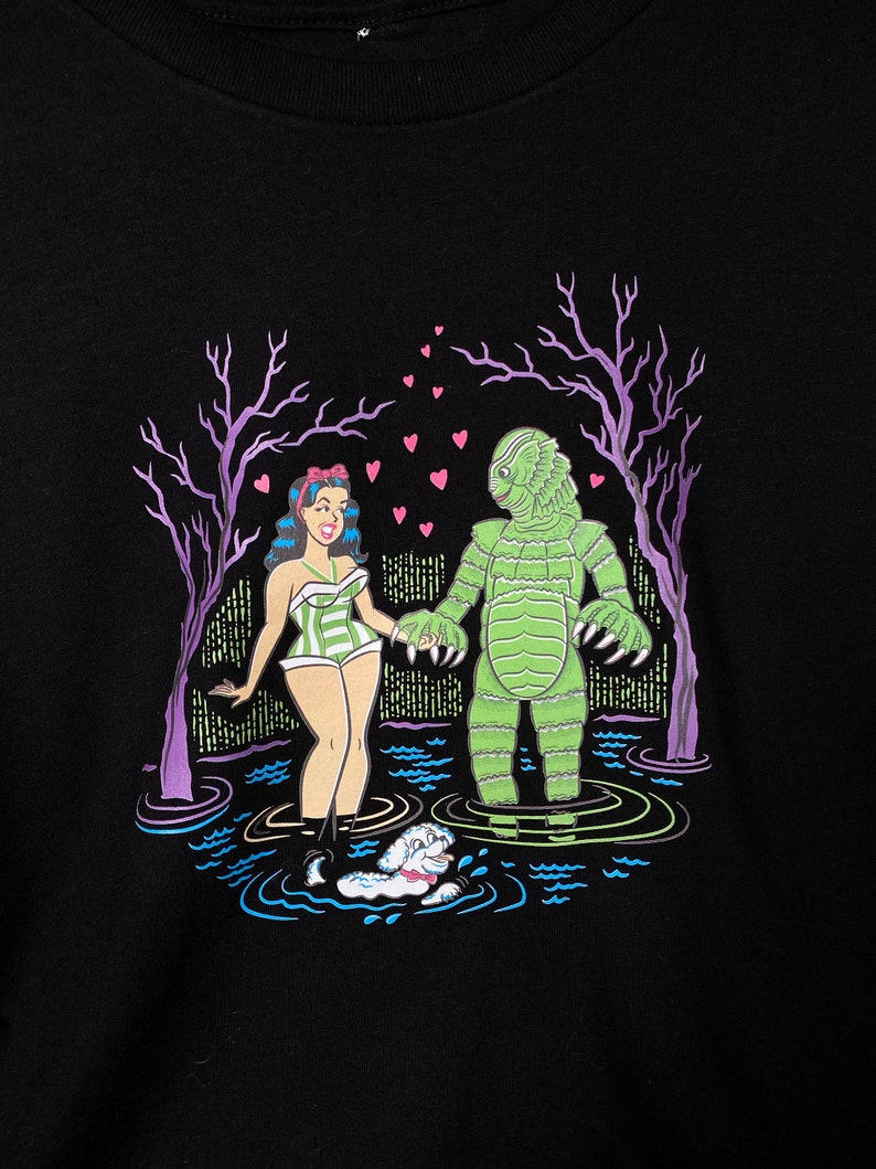 May include: Black graphic t-shirt featuring a cartoon illustration of a woman in a striped swimsuit holding hands with a green monster in a swamp. Pink hearts float above them, with a small dog swimming in the water. Purple trees frame the scene.