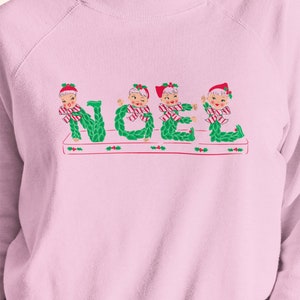 Puede incluir: Una sudadera rosa con un diseño navideño que presenta cuatro elfos formando la palabra "NOEL" con sus cuerpos. Los elfos llevan gorros rojos, camisas a rayas verdes y blancas y lazos verdes.