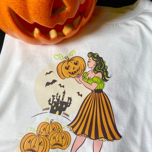 May include: A white t-shirt with a vintage-style illustration of a woman holding a jack-o'-lantern. The woman is wearing a green and yellow dress with a black and yellow striped skirt. The background features a castle and bats.