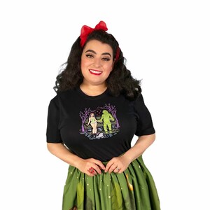 May include: Black t-shirt featuring a graphic print of a creature from the black lagoon and a woman holding hands in a swamp. The design is in shades of green, purple, and pink. The shirt is made of a soft, comfortable material.
