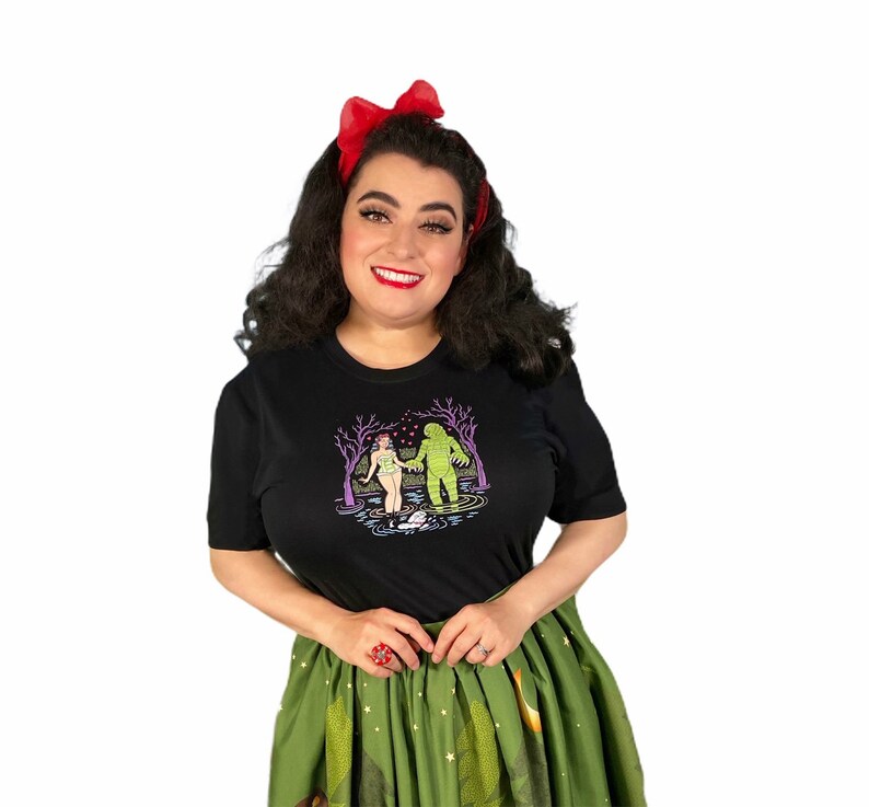 May include: Black t-shirt featuring a graphic print of a creature from the black lagoon and a woman holding hands in a swamp. The design is in shades of green, purple, and pink. The shirt is made of a soft, comfortable material.