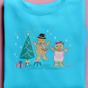 Puede incluir: Una sudadera azul turquesa con un diseño navideño que presenta a dos personas de jengibre decorando un árbol de Navidad con luces de colores. Las personas de jengibre sonríen y visten atuendos festivos.