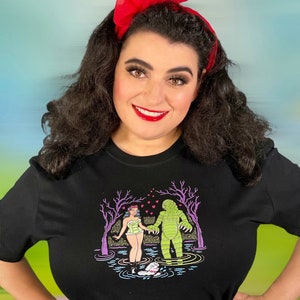 May include: Black t-shirt featuring a colorful graphic of a creature from the black lagoon holding hands with a woman in a swimsuit. The woman is wearing a floral crown. The background is a colorful swamp scene.
