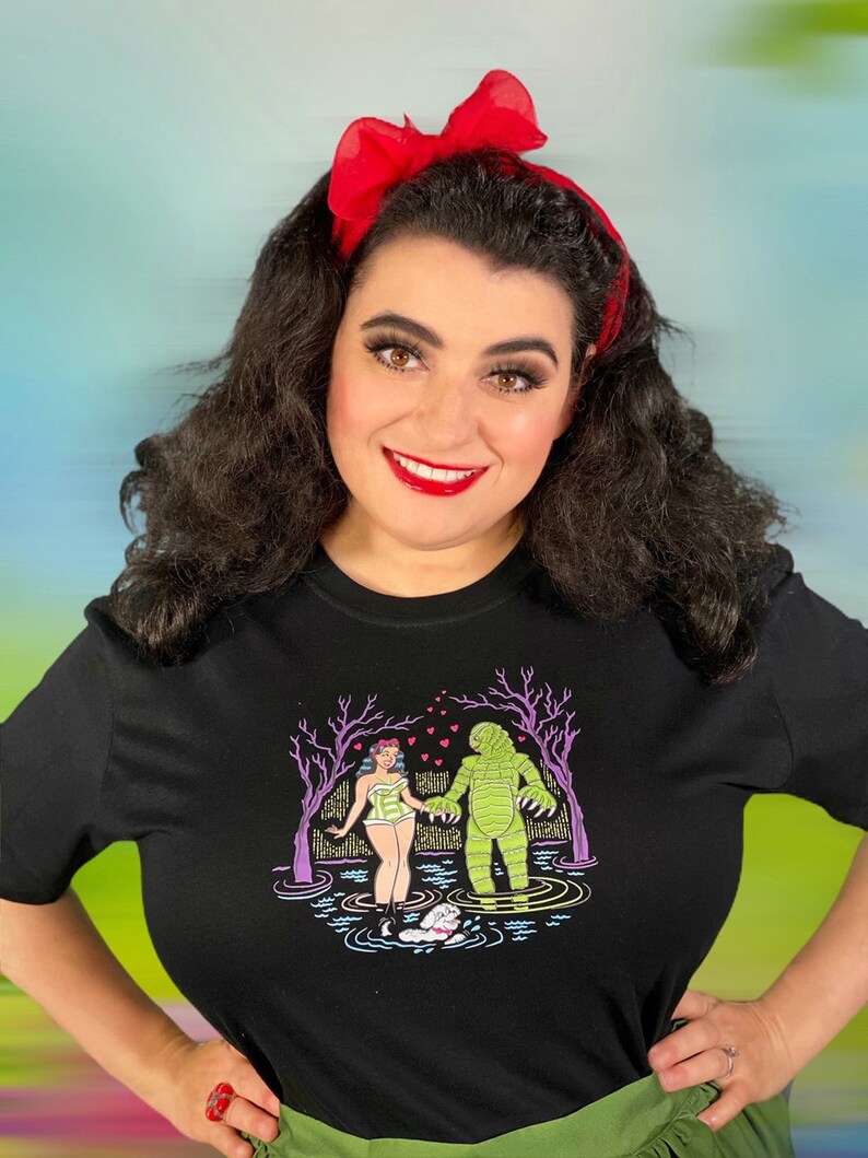 May include: Black t-shirt featuring a colorful graphic of a creature from the black lagoon holding hands with a woman in a swimsuit. The woman is wearing a floral crown. The background is a colorful swamp scene.