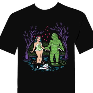 May include: Black t-shirt with a graphic print of a woman in a green and white swimsuit holding hands with a green monster. The monster is standing in water and there are hearts floating around them.
