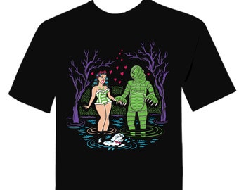 Könnte beinhalten: Schwarzes T-Shirt mit einem Grafikdruck einer Frau in einem grün-weißen Badeanzug, die mit einem grünen Monster Händchen hält. Das Monster steht im Wasser und es schweben Herzen um sie herum.
