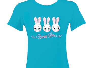 Peut inclure: Un t-shirt bleu turquoise avec un motif de trois lapins blancs avec des nez et des yeux roses. Les lapins ont des expressions différentes. Le texte "Bunny Love" est écrit en rose sous les lapins.