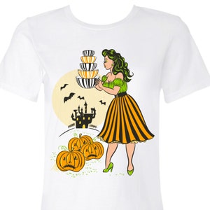 Puede incluir: Camiseta blanca con un diseño retro de Halloween. El gráfico incluye una mujer sosteniendo cuencos apilados, calabazas, murciélagos, un castillo y una luna llena. La mujer lleva un top verde, una falda a rayas naranja y negra y tacones verdes.