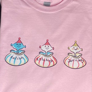 Puede incluir: Camiseta rosa con un gráfico de tres tarros de dibujos animados con caras. Los tarros están etiquetados como "Relish", "Ketchup" y "Jam 'n Jelly".
