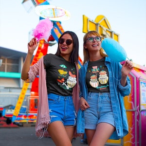 May include: Two friends wearing black t-shirts with colorful graphic prints. The t-shirts feature illustrations of jars with the text "Sugar & Spice" and "Everything Nice". The friends are holding pink and blue cotton candy.