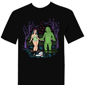 May include: Black t-shirt featuring a cartoon illustration of a woman in a green and white swimsuit holding hands with a green monster in a swamp. A small white dog swims nearby, with purple trees and pink hearts in the background.