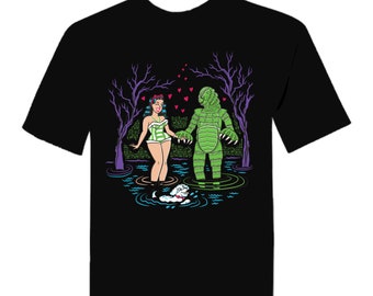 Könnte beinhalten: Schwarzes T-Shirt mit einer Cartoon-Illustration einer Frau in einem grün-weißen Badeanzug, die die Hand eines grünen Monsters in einem Sumpf hält. Ein kleiner weißer Hund schwimmt in der Nähe, mit lila Bäumen und rosa Herzen im Hintergrund.