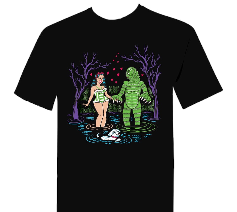 May include: Black t-shirt featuring a cartoon illustration of a woman in a green and white swimsuit holding hands with a green monster in a swamp. A small white dog swims nearby, with purple trees and pink hearts in the background.