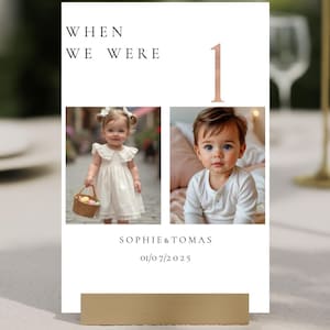 以下が含まれることがあります： 「WHEN WE WERE 1」の文字と2枚の赤ちゃんの写真が載った白いカード。カードには「SOPHIE&TOMAS」と日付「01/07/2025」も記載されています。金色のスタンドがカードを支えています。