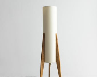 Könnte beinhalten: Ein weißer zylindrischer Lampenschirm mit einem dreibeinigen Holzfuß. Der Lampenschirm ist aus Stoff und der Fuß aus Holz. Die Lampe hat ein Kabel mit einem Schalter.