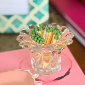 May include: A clear glass match holder with a scalloped rim containing a cluster of wooden matches with sage green tips. The small, transparent vessel features vertical fluted detailing along its base, providing a sturdy and decorative container.