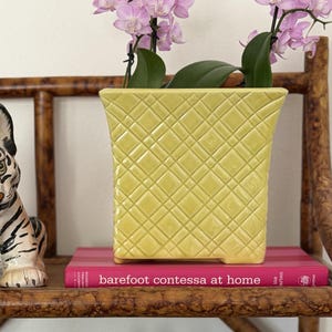 May include: A brown faux bamboo shelf holding a ceramic tiger figurine, a yellow lattice-patterned planter with pink orchids, a Barefoot Contessa at Home book, a green lidded jar, and two yellow Barefoot Contessa cookbooks.