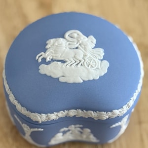 Może przedstawiać: Małe, okrągłe, niebieskie pudełko na drobiazgi z białym reliefem przedstawiającym rydwan i konie. Pudełko ma białe, dekoracyjne obramowanie. Pudełko stoi na drewnianej powierzchni.