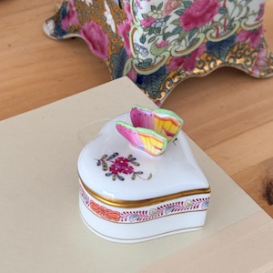 May include: A heart-shaped porcelain trinket box with a butterfly lid. The box is white with gold trim and floral designs. A vase with pink roses and a decorative gold framed object are in the background.