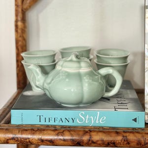 Puede incluir: Una tetera de cerámica verde claro con forma de calabaza y detalles florales en relieve, acompañada de cuatro tazas de té pequeñas a juego. El conjunto está dispuesto sobre un libro titulado Tiffany Style con lomo de color turquesa.