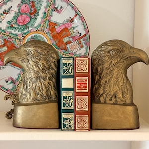 May include: Two brass eagle head bookends flank two antique books with green and red covers. A decorative plate with a floral and figural design is in the background. The bookends and books are on a white shelf.