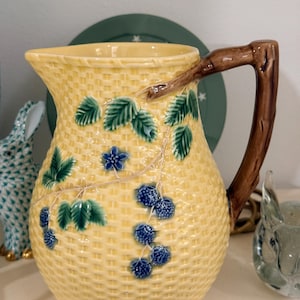 May include: Yellow ceramic pitcher with a woven basket design. The pitcher features a brown branch handle and is decorated with green leaves and blue berries. A decorative piece for serving beverages.