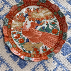 Puede incluir: Un plato decorativo con borde festoneado con un vibrante diseño de pavo real. El plato muestra un pavo real rojo y naranja con sus plumas extendidas, rodeado de elementos florales y follaje en rojo, verde y dorado. El borde tiene un patrón detallado.