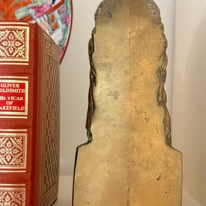 May include: A vintage brass bookend in the shape of a bust. The bookend has a flat back and a detailed front with braided hair. The bookend is placed on a white shelf next to a red book with gold lettering and a decorative plate.