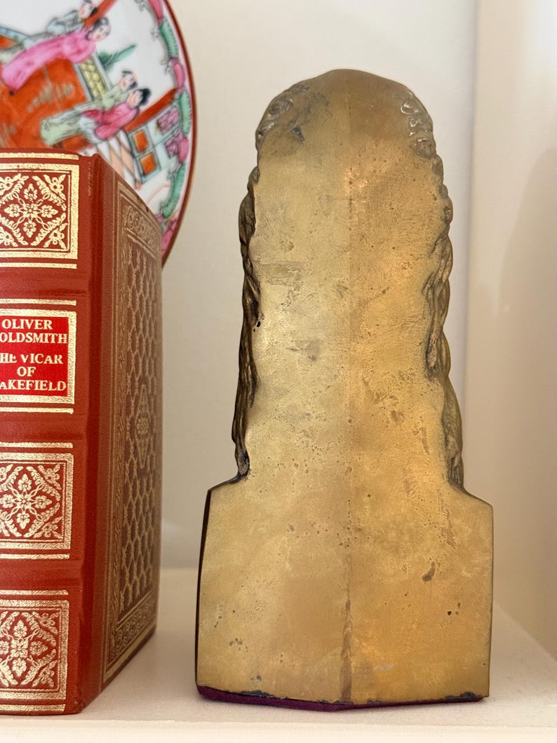 May include: A vintage brass bookend in the shape of a bust. The bookend has a flat back and a detailed front with braided hair. The bookend is placed on a white shelf next to a red book with gold lettering and a decorative plate.