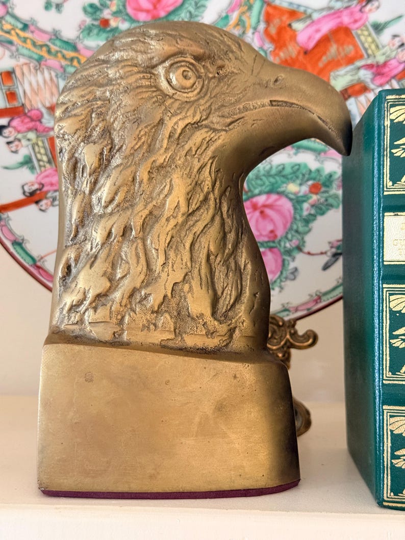 May include: A brass eagle head bookend. The bookend is a detailed sculpture of an eagle's head and beak, with textured feathers. The base is rectangular. The bookend is a decorative item for home decor.