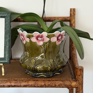 Puede incluir: Una maceta de cerámica verde oliva brillante con borde festoneado y relieves de flores rosas con centros oscuros y follaje verde. El recipiente tiene un cuerpo ancho y curvado con una base ondulada, conteniendo una planta de orquídea de hojas largas.