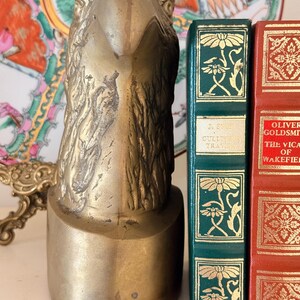 May include: A brass eagle head bookend with a detailed, textured design. The bookend is positioned next to two antique books with decorative covers, one green and one red. A decorative plate is visible in the background.