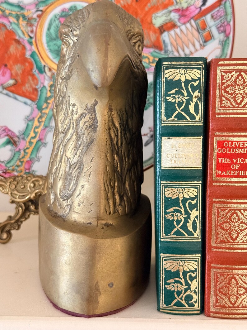 May include: A brass eagle head bookend with a detailed, textured design. The bookend is positioned next to two antique books with decorative covers, one green and one red. A decorative plate is visible in the background.