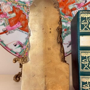 May include: A gold-colored, vintage bookend with a detailed, sculpted design. The bookend has a rectangular shape with a flat base and a decorative top. A green book with gold accents is visible in the background.