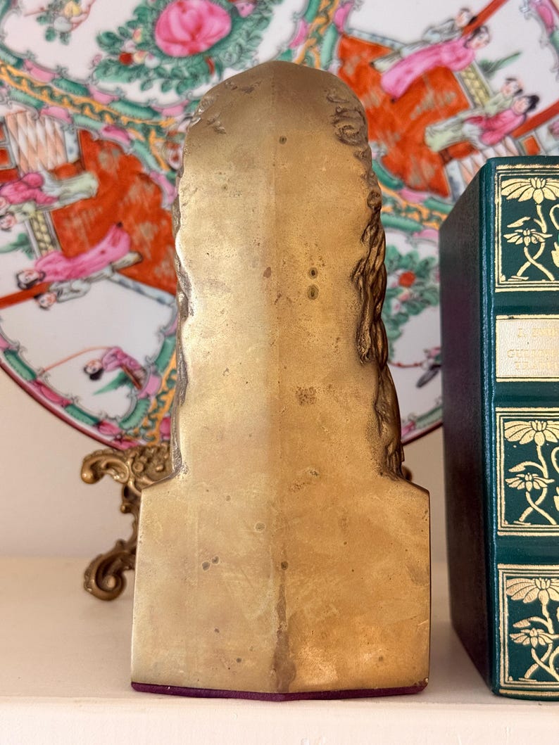 May include: A gold-colored, vintage bookend with a detailed, sculpted design. The bookend has a rectangular shape with a flat base and a decorative top. A green book with gold accents is visible in the background.