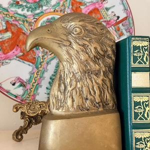 May include: A brass eagle head bookend with detailed feather texture. The bookend has a rectangular base with a red felt bottom. A green book with gold floral accents is next to the bookend. A decorative plate with pink and green floral designs is in the background.