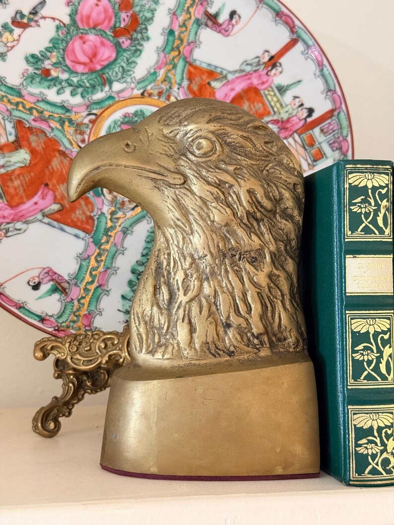 May include: A brass eagle head bookend with detailed feather texture. The bookend has a rectangular base with a red felt bottom. A green book with gold floral accents is next to the bookend. A decorative plate with pink and green floral designs is in the background.