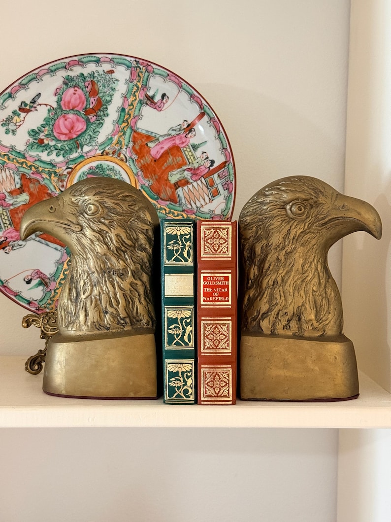 May include: Two brass eagle head bookends flank two antique books with green and red covers. A decorative plate with a floral and figural design is in the background. The bookends and books are on a white shelf.