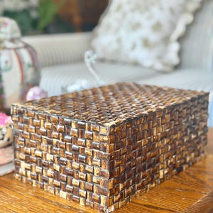 May include: A rectangular storage box featuring a woven exterior pattern made of dark brown and tan material. The box has a flat lid and a structured shape, displaying a consistent grid-like texture across all visible sides and the top surface.