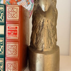 May include: A brass eagle head sculpture with detailed feather texture, mounted on a cylindrical base. The sculpture is displayed next to a red book with gold lettering and a decorative plate.