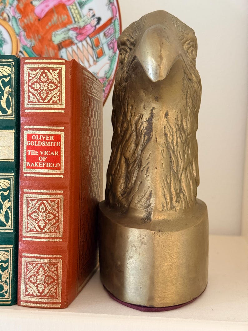 May include: A brass eagle head sculpture with detailed feather texture, mounted on a cylindrical base. The sculpture is displayed next to a red book with gold lettering and a decorative plate.