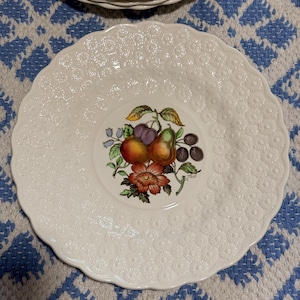 May include: White ceramic plate with a textured surface and a scalloped edge. The center features a colorful illustration of fruit, including pears, plums, and a flower. A stack of similar plates is visible in the background.