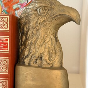 May include: A golden-colored eagle head bookend. The bookend is made of metal and has a detailed texture. The eagle's head is in profile, with a strong beak and focused eye. The bookend is on a shelf next to a red book.