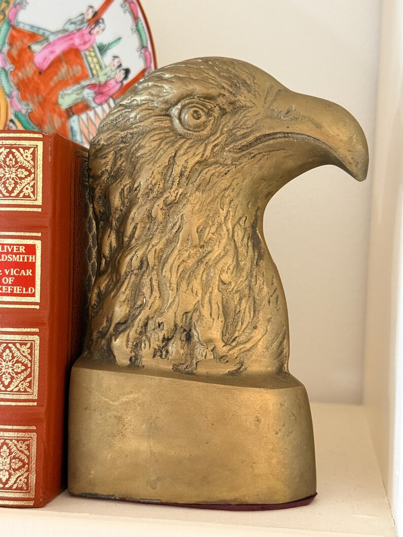 May include: A golden-colored eagle head bookend. The bookend is made of metal and has a detailed texture. The eagle's head is in profile, with a strong beak and focused eye. The bookend is on a shelf next to a red book.