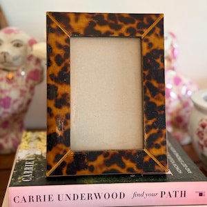 May include: A rectangular picture frame with a brown and black tortoiseshell pattern. The frame has a smooth, glossy finish and mitered corners. It is positioned vertically, featuring a plain, light-coloured insert within the central opening.