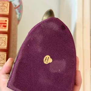 May include: A decorative, purple velvet object with a gold-colored metal top. The base has a gold trim and a small, round sticker that reads "MADE IN KOREA". The object is held in a person's hand.