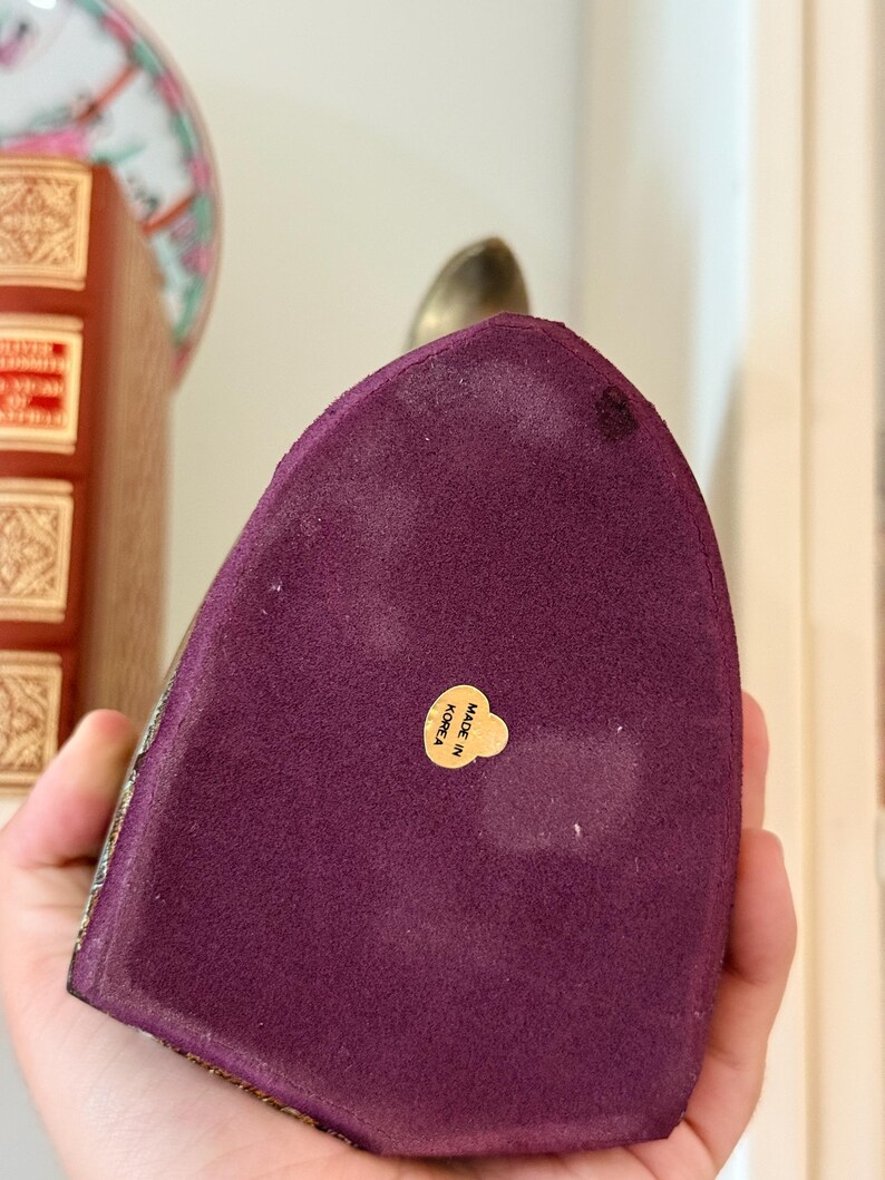 May include: A decorative, purple velvet object with a gold-colored metal top. The base has a gold trim and a small, round sticker that reads "MADE IN KOREA". The object is held in a person's hand.